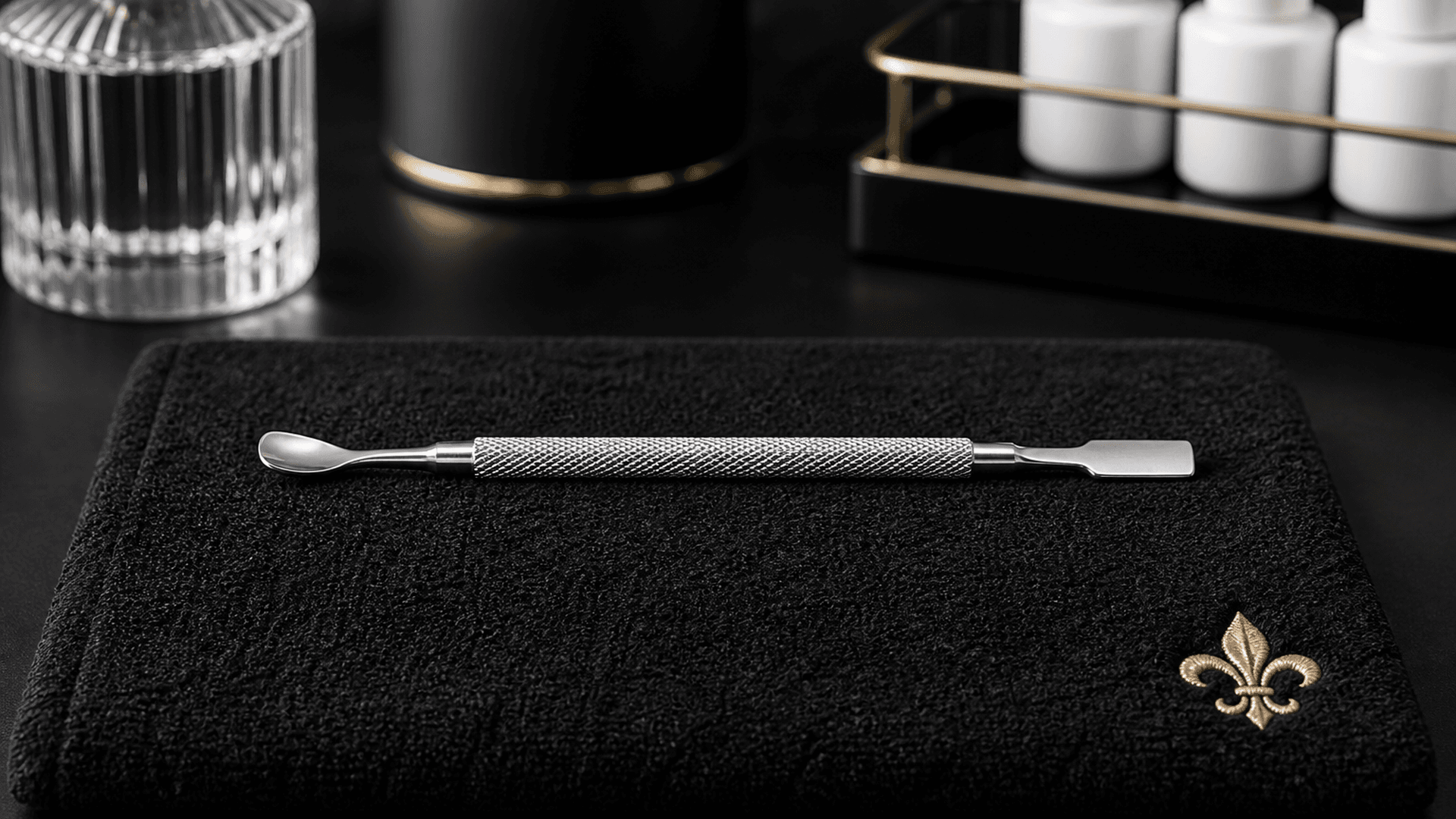 Single stainless dual-ended cuticle pusher on a matte black manicure station with brushed gold fleur accent — Lou Essentials